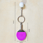 A table tennis-themed keychain with a white ball charm, silver metal chain and ring, and a pink paddle-shaped charm on a beige textured background with dimension lines and text.