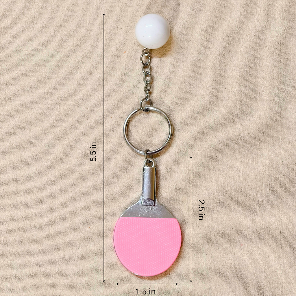 A close-up of pink ping-pong paddle keychain charm and a white ball accessory hanging from a gold-tone ring, showing measurements in the beige background.