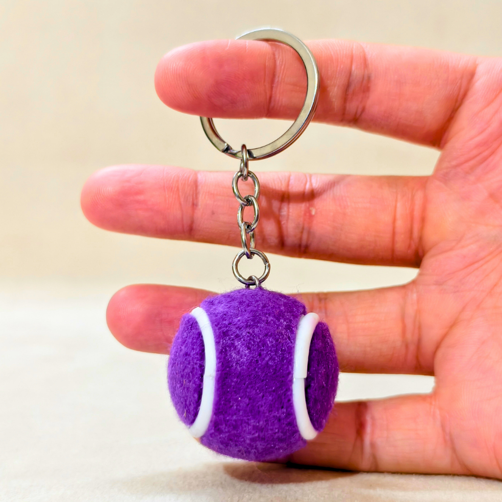 A purple tennis ball keychain charm with white curved stripes hangs from a silver metal key ring held in hand on the beige background 