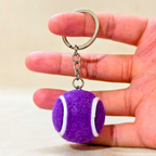 A purple tennis ball keychain charm with white curved stripes hangs from a silver metal key ring held in hand on the beige background 