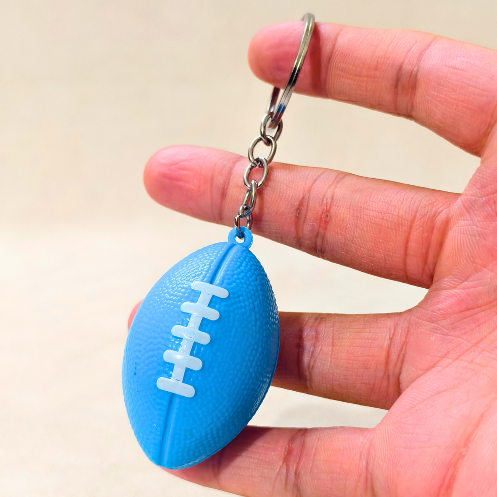A small blue football-shaped keychain with white laces and a silver key ring, held between a person's fingers against a light beige background.