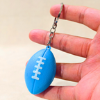 A small blue football-shaped keychain with white laces and a silver key ring, held between a person's fingers against a light beige background.