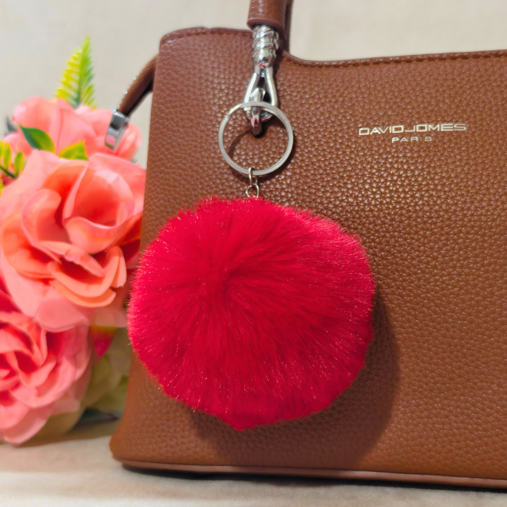 Red pom-pom keychain attached to a brown handbag with flowers in the background