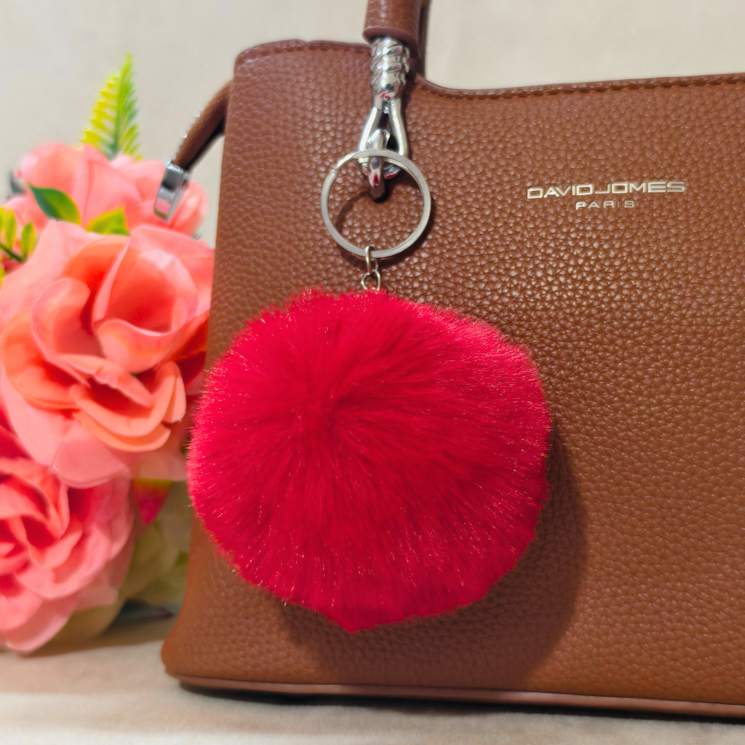 Red pom-pom keychain attached to a brown handbag with flowers in the background