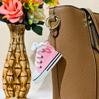 A miniature pink canvas high-top sneaker keychain charm with white laces and black stripes, attached to a gold key ring on a brown textured handbag, with a decorative floral vase in the background.