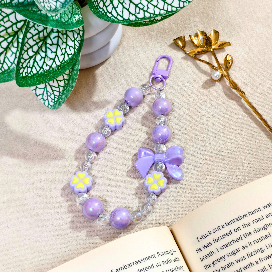 Purple beaded keychain with flowers on a beige surface with an open book and decorative elements.