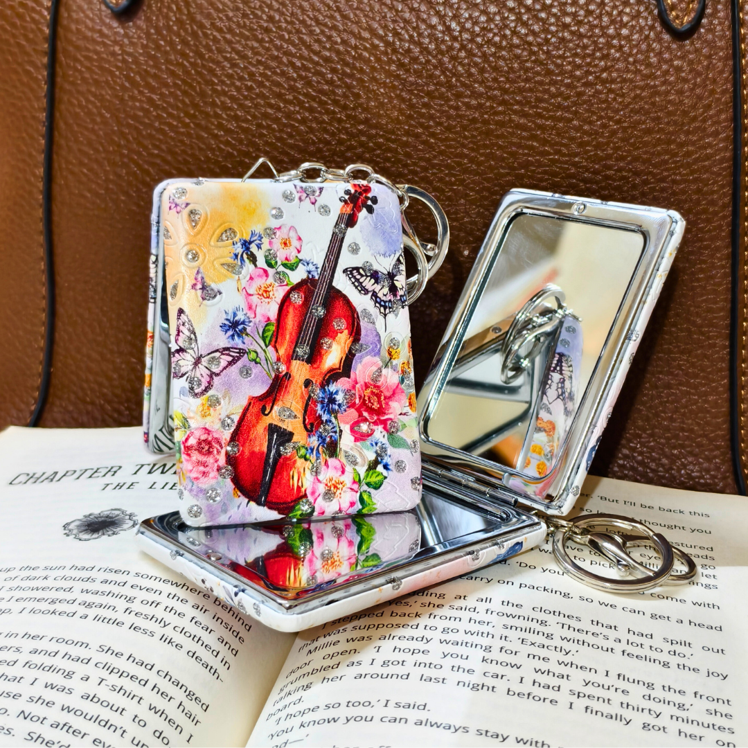 Compact mirror with floral and violin design on a book next to a keychain.
