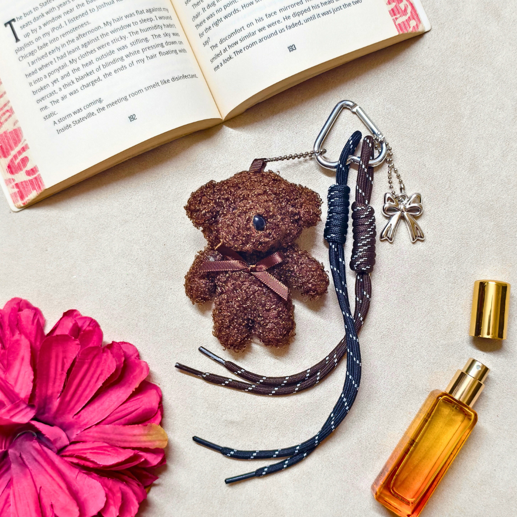Brown teddy bear keychain, braided keychain, and perfume bottle on a textured surface with an open book and pink flower.