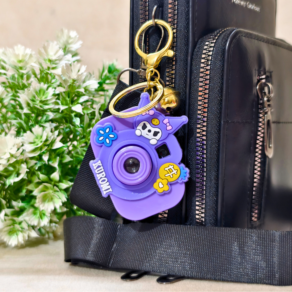 Purple camera-shaped keychain attached to a black bag with greenery in the background