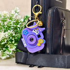 Purple camera-shaped keychain attached to a black bag with greenery in the background