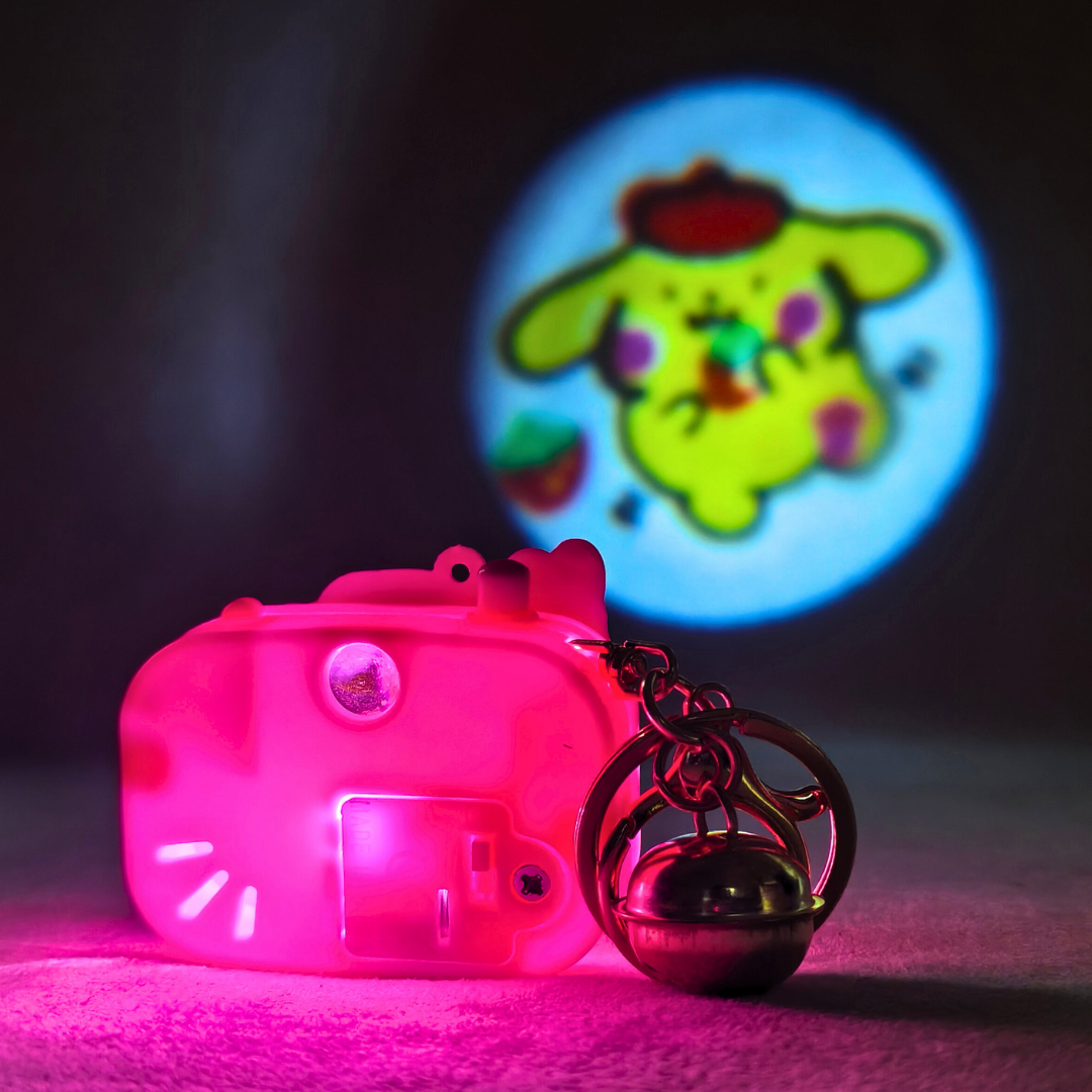 Pink camera-shaped object with a bell charm in front of a colorful cartoon character projection.