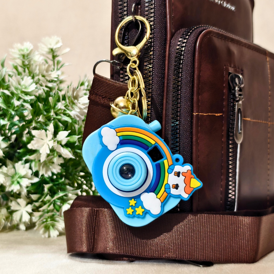A colorful camera-shaped keychain charm with a rainbow, clouds, stars, and a small dog character, attached to a gold clasp and hanging on a brown backpack. A blurred plant is visible in the background.