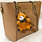 A tan pebbled leather tote bag with gold-tone hardware and a hanging plush teddy bear keychain. The bag has black trim and a small gold logo reading 'FASHION & MADE' on the front.
