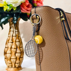 A tennis-themed keychain featuring a miniature silver metal tennis racket and a yellow tennis ball charm, shown hanging from a brown textured leather handbag next to a decorative vase with flowers.