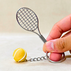 A miniature metal tennis racket keychain with a small yellow tennis ball charm, held by a hand against a neutral beige background.