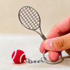 A miniature silver metal tennis racket keychain with a small red and white felt tennis ball charm, held by a hand against a neutral beige background.