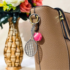 A silver tennis racket keychain charm with a pink tennis ball accent hanging from a tan textured leather handbag, with a decorative vase of flowers in the background.