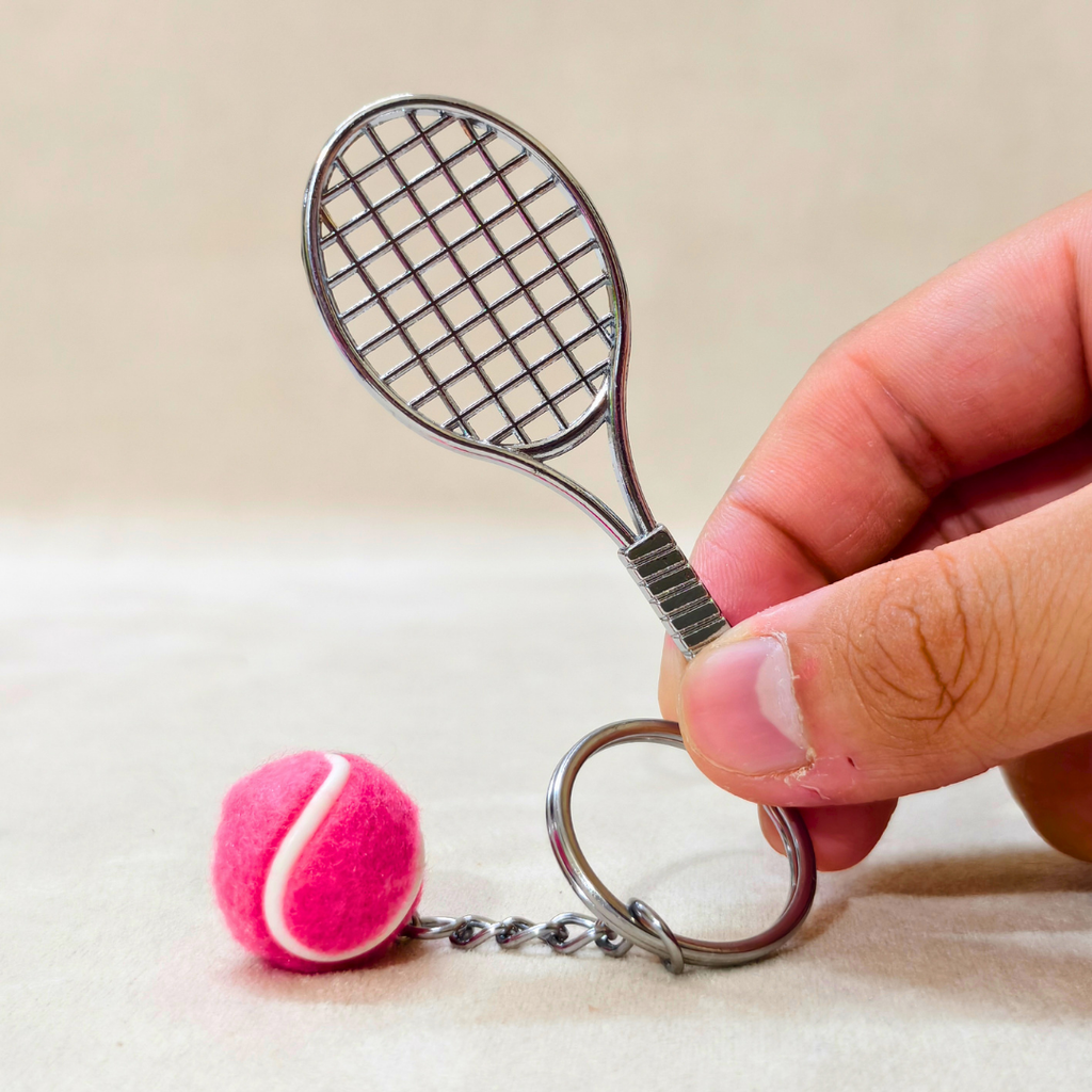 A miniature metal tennis racket keychain with a pink felt tennis ball charm, held by a hand against a neutral beige background.