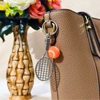 A tennis-themed keychain charm featuring a silver metal tennis racket and an orange tennis ball, attached to a tan textured leather handbag. A decorative ceramic vase with flowers is blurred in the background.