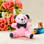 A pink plush teddy bear with black polka-dot bow tie and black-and-white polka-dot feet, styled on a beige surface with artificial pink roses and a blue perfume bottle in the background.