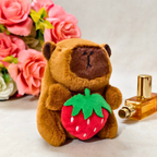Small brown bear plush toy holding a strawberry next to a perfume bottle and flowers.