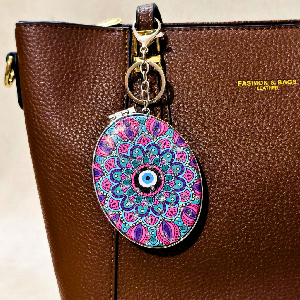 A brown pebbled leather handbag with a colorful oval charm featuring a vibrant mandala pattern and a central evil eye design, attached to a gold-tone key ring.