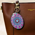 A brown pebbled leather handbag with a colorful oval charm featuring a vibrant mandala pattern and a central evil eye design, attached to a gold-tone key ring.