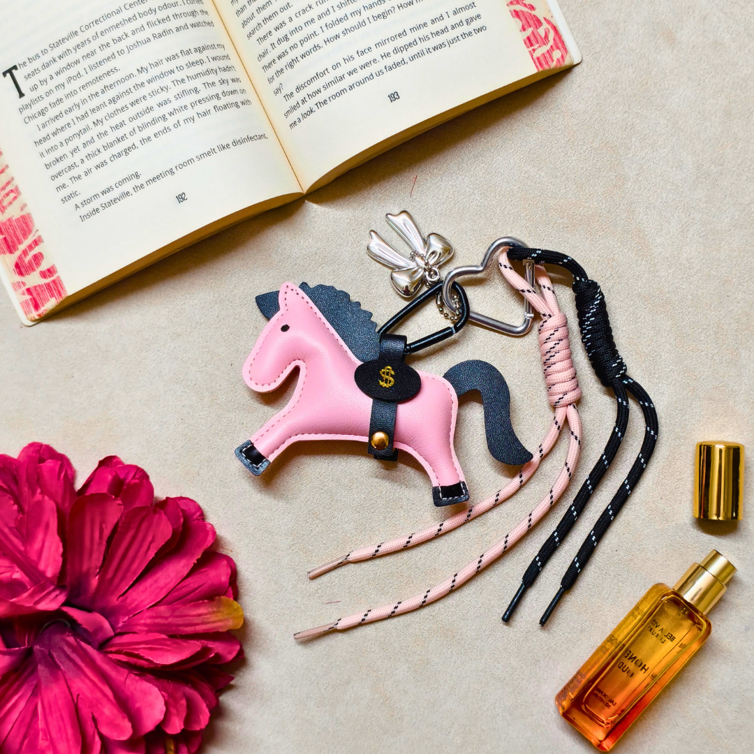 Pink horse-shaped keychain with braided straps on a beige surface with a book and flower.