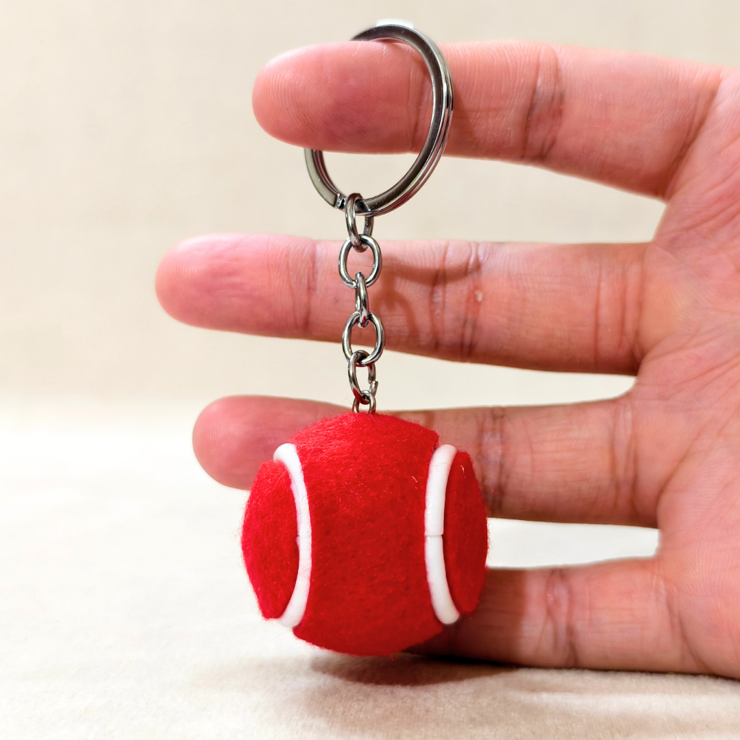 A red felt tennis ball keychain with white stitched lines and a silver metal key ring, held in a person's hand against a neutral beige background.