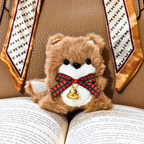 A small brown plush teddy bear toy with a white belly, black eyes, and a red plaid bow tie with a gold bell, resting on an open book with patterned neckties in the background.