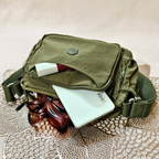 Military Green Sling Bag With Items In Front Of Beige Background.
