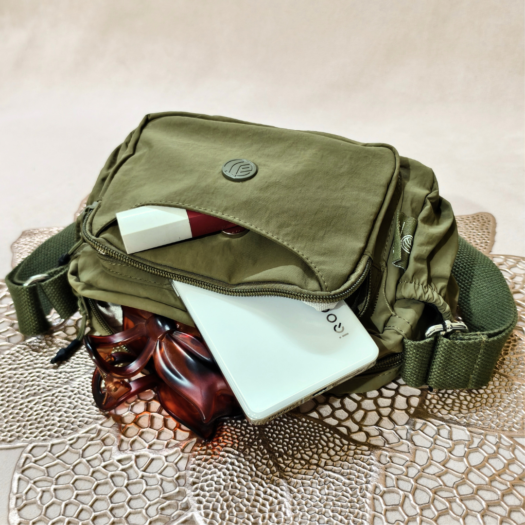 Military Green Sling Bag With Items In Front Of Beige Background.