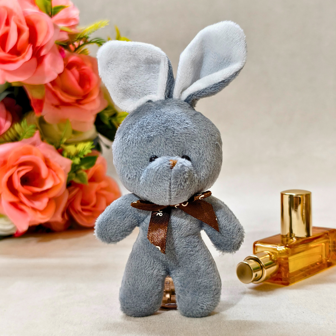 Gray plush bunny with a brown bow tie next to a perfume bottle and pink flowers.
