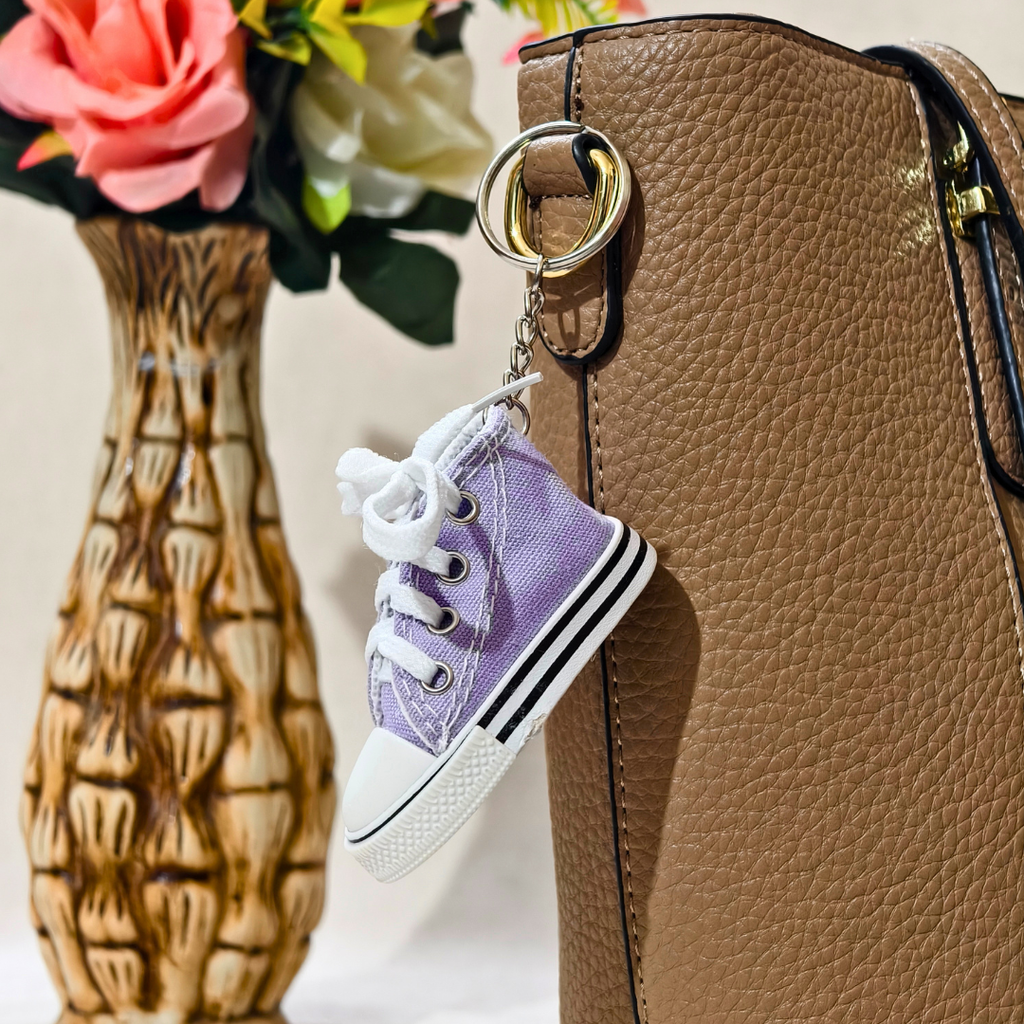 A miniature lavender purple canvas high-top sneaker keychain with white laces and a gold metal ring, shown hanging from a brown textured handbag next to a decorative vase of flowers.
