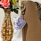 A miniature lavender purple canvas high-top sneaker keychain with white laces and a gold metal ring, shown hanging from a brown textured handbag next to a decorative vase of flowers.
