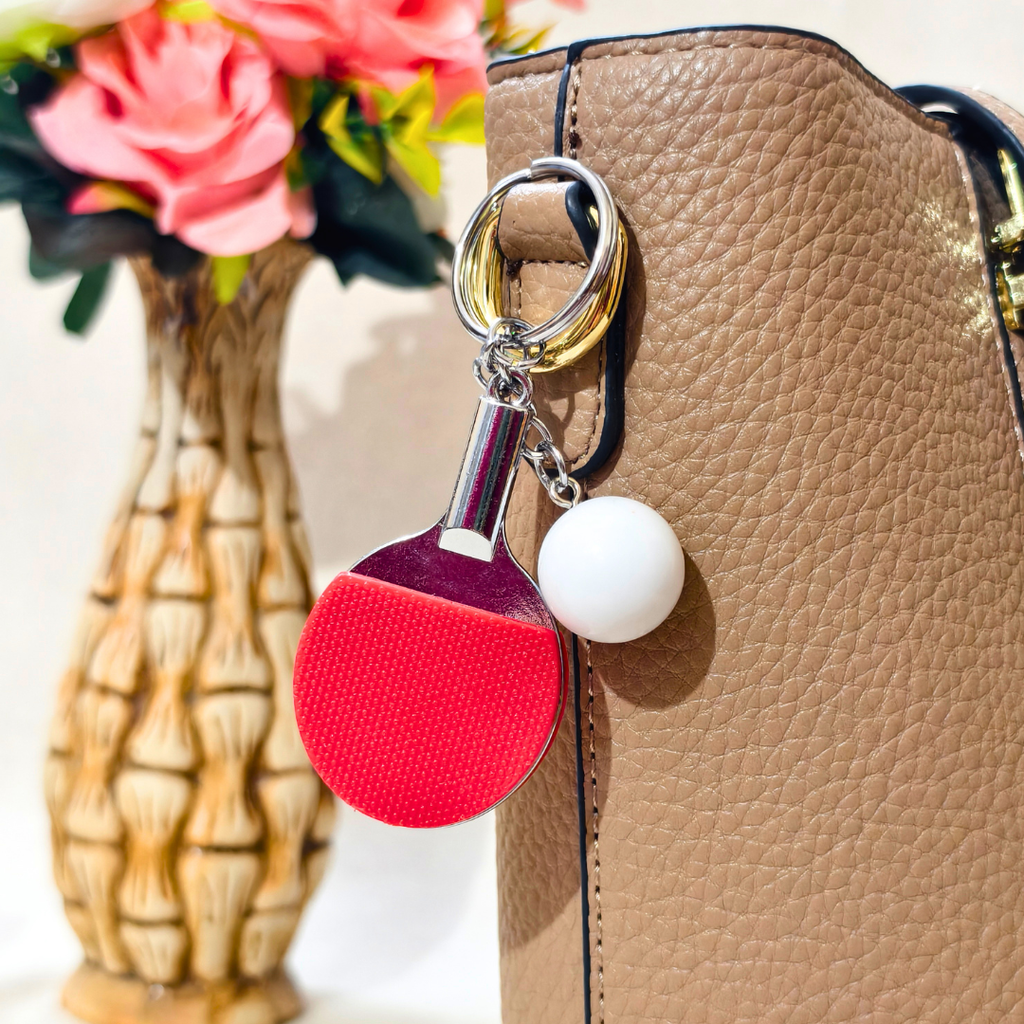 A close-up of a tan textured handbag with a decorative keychain charm shaped like a ping-pong paddle and a white ball, with a blurred vase of pink flowers in the background.