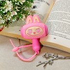 A pink bunny-shaped hair clip with a cute face and a matching pink beaded wrist chain, styled on an open book with white flowers in the corner.