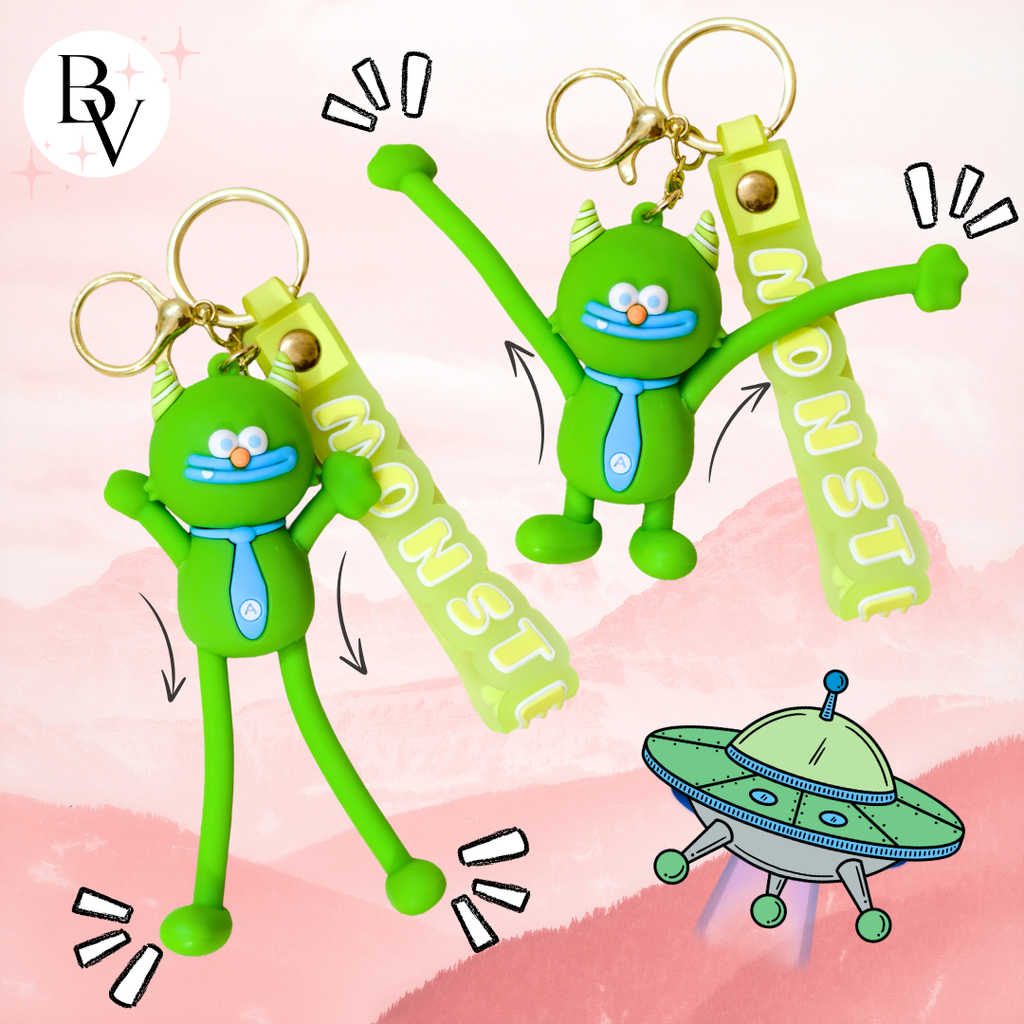 Two green cartoon monster keychain charms with long limbs and gold keyrings, shown on a pink illustrated background with a UFO graphic and a visible BV logo in the top left.