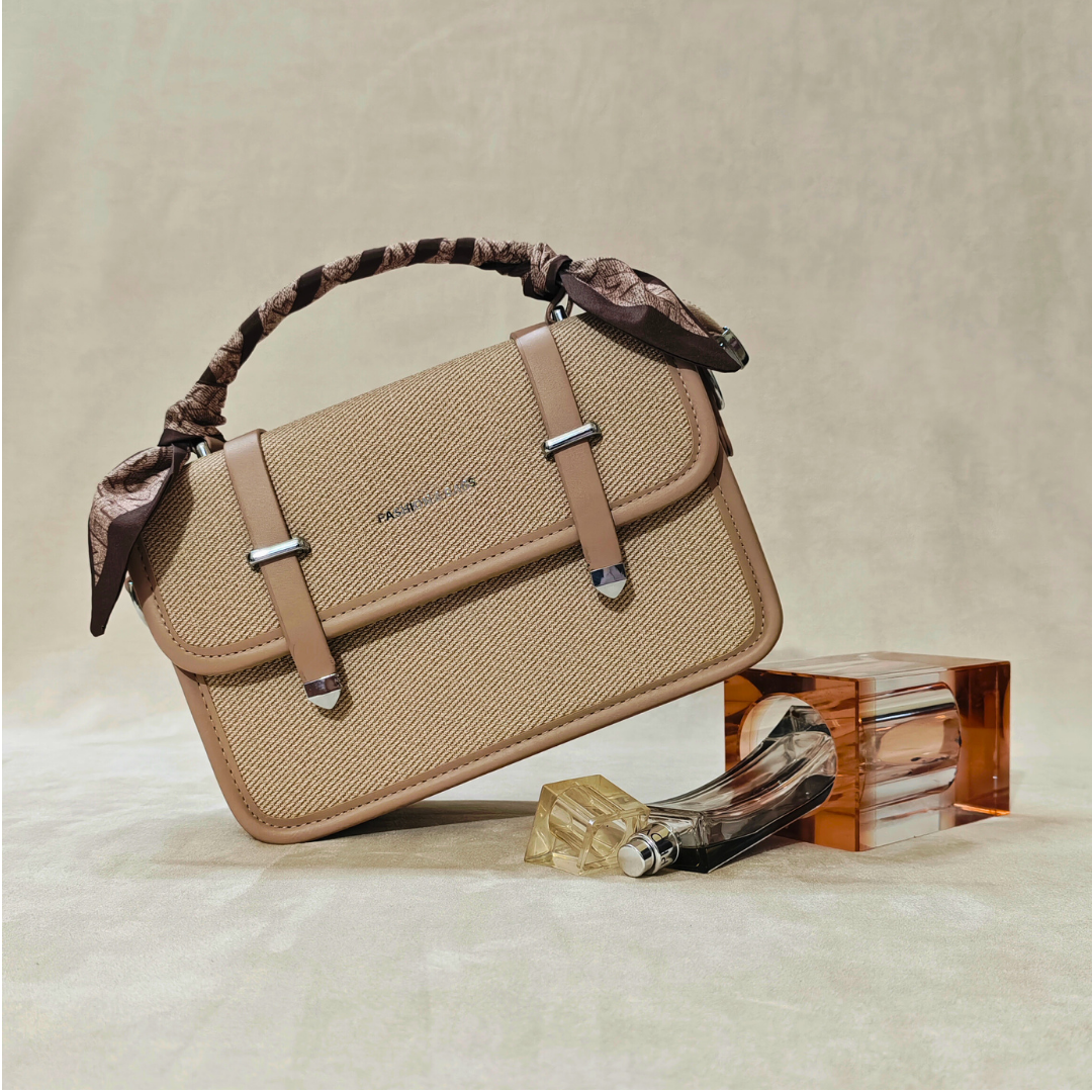 Beige handbag with decorative handles on a textured surface with a bottle and box.