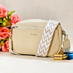 A beige faux-leather crossbody bag with a pebbled texture, featuring a wide beige and white chevron-patterned strap and the brand name 'Beauty' printed on the front. The bag is styled with pink flowers and a perfume bottle on a light beige surface.