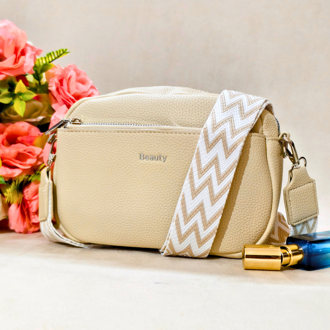A beige faux-leather crossbody bag with a pebbled texture, featuring a wide beige and white chevron-patterned strap and the brand name 'Beauty' printed on the front. The bag is styled with pink flowers and a perfume bottle on a light beige surface.
