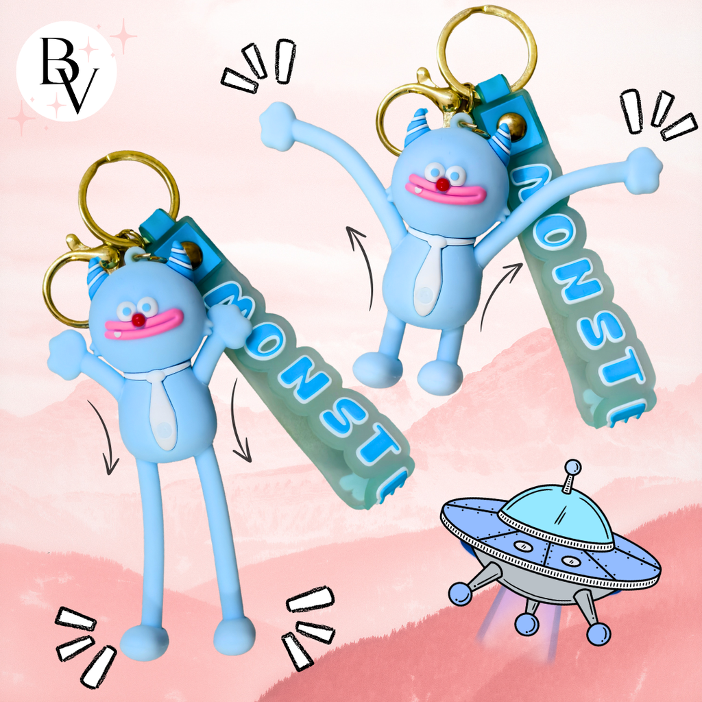 Two light blue monster-shaped keychain charms with gold keyrings and translucent green tags reading MONSTER, shown on a pink illustrated background with a cartoon UFO.