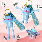 Two light blue monster-shaped keychain charms with gold keyrings and translucent green tags reading MONSTER, shown on a pink illustrated background with a cartoon UFO.