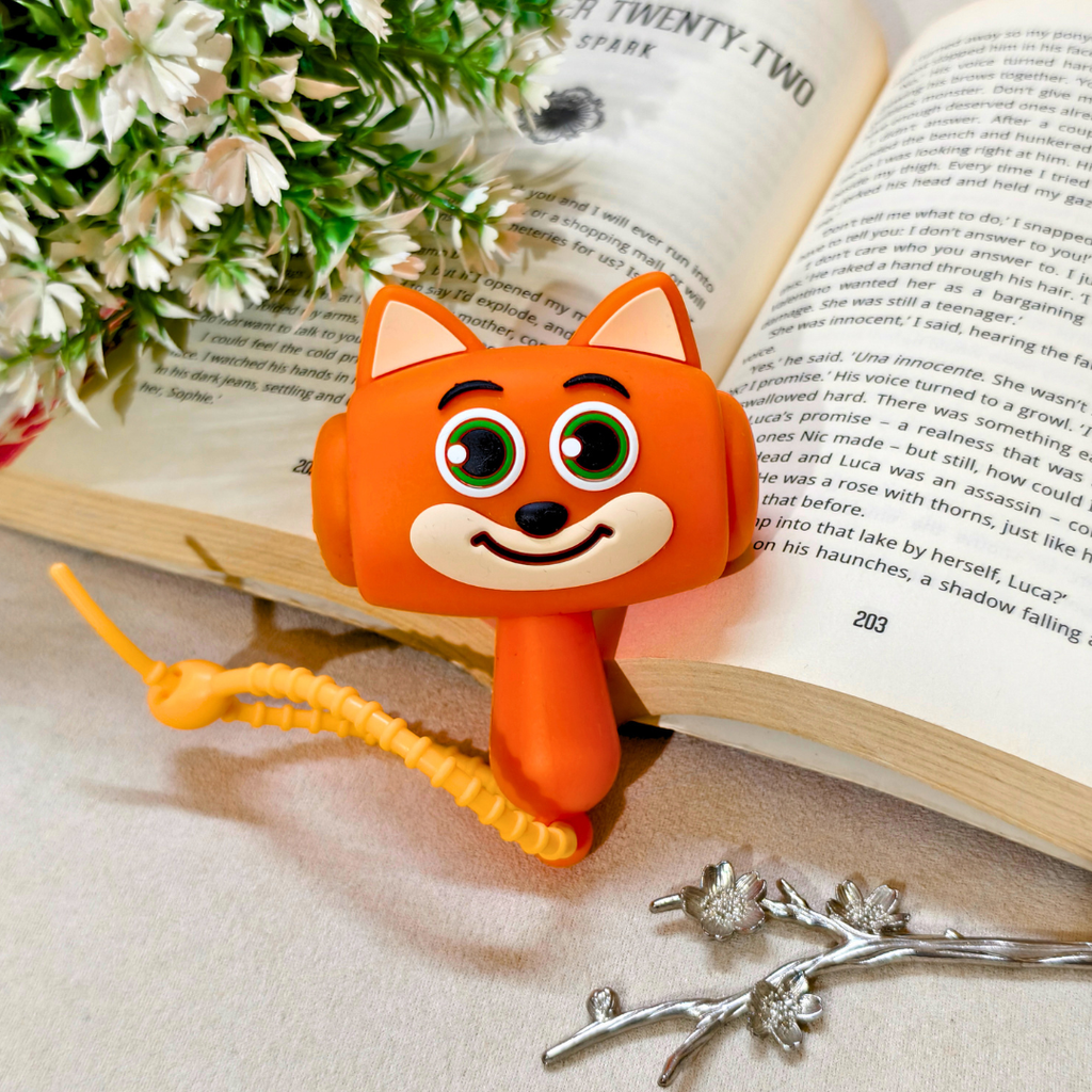 An orange fox-shaped bookend with large green eyes and a smiling face, holding an open book. The book is styled with white flowers and a silver branch on a beige surface.