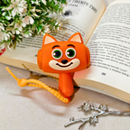 An orange fox-shaped bookend with large green eyes and a smiling face, holding an open book. The book is styled with white flowers and a silver branch on a beige surface.