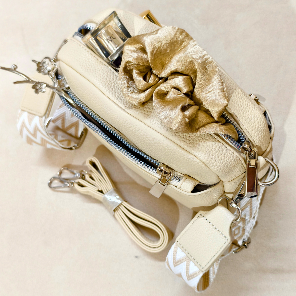 Cream-colored faux-leather crossbody bag with a gold satin scrunchie detail, silver zippers, and a patterned cream-and-white strap, displayed on a neutral beige background.