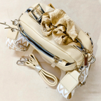 Cream-colored faux-leather crossbody bag with a gold satin scrunchie detail, silver zippers, and a patterned cream-and-white strap, displayed on a neutral beige background.