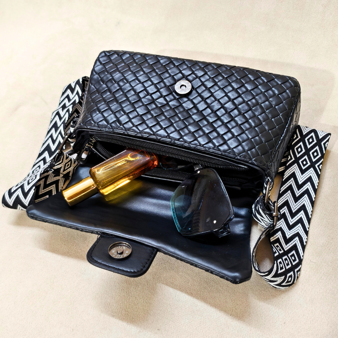 Black woven handbag with a patterned strap, containing a bottle and sunglasses on a beige background.