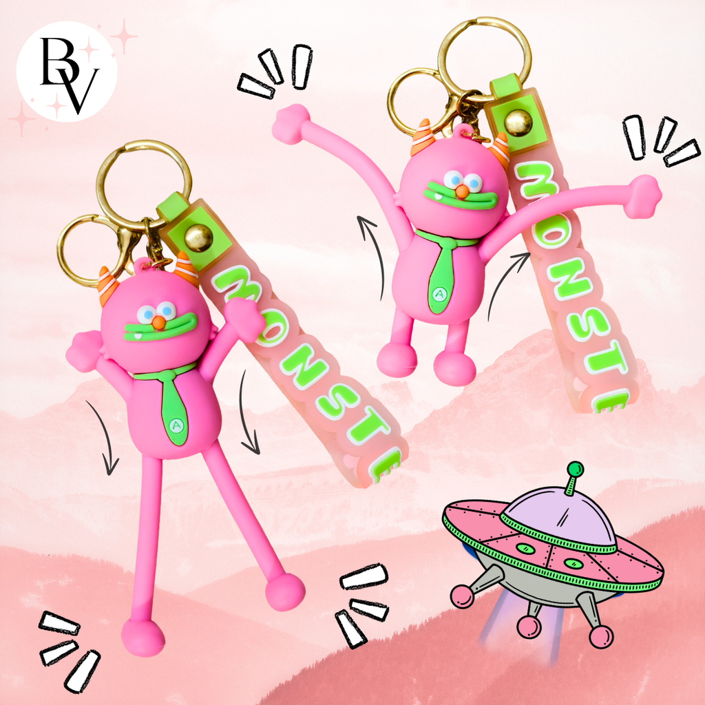 Two pink monster-shaped keychains with green neckties and gold keyrings, each holding a translucent pink tag that reads MONSTIE. The background is a soft pink illustrated landscape with cartoon sound effects and a small UFO graphic.