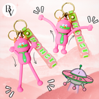 Two pink monster-shaped keychains with green neckties and gold keyrings, each holding a translucent pink tag that reads MONSTIE. The background is a soft pink illustrated landscape with cartoon sound effects and a small UFO graphic.